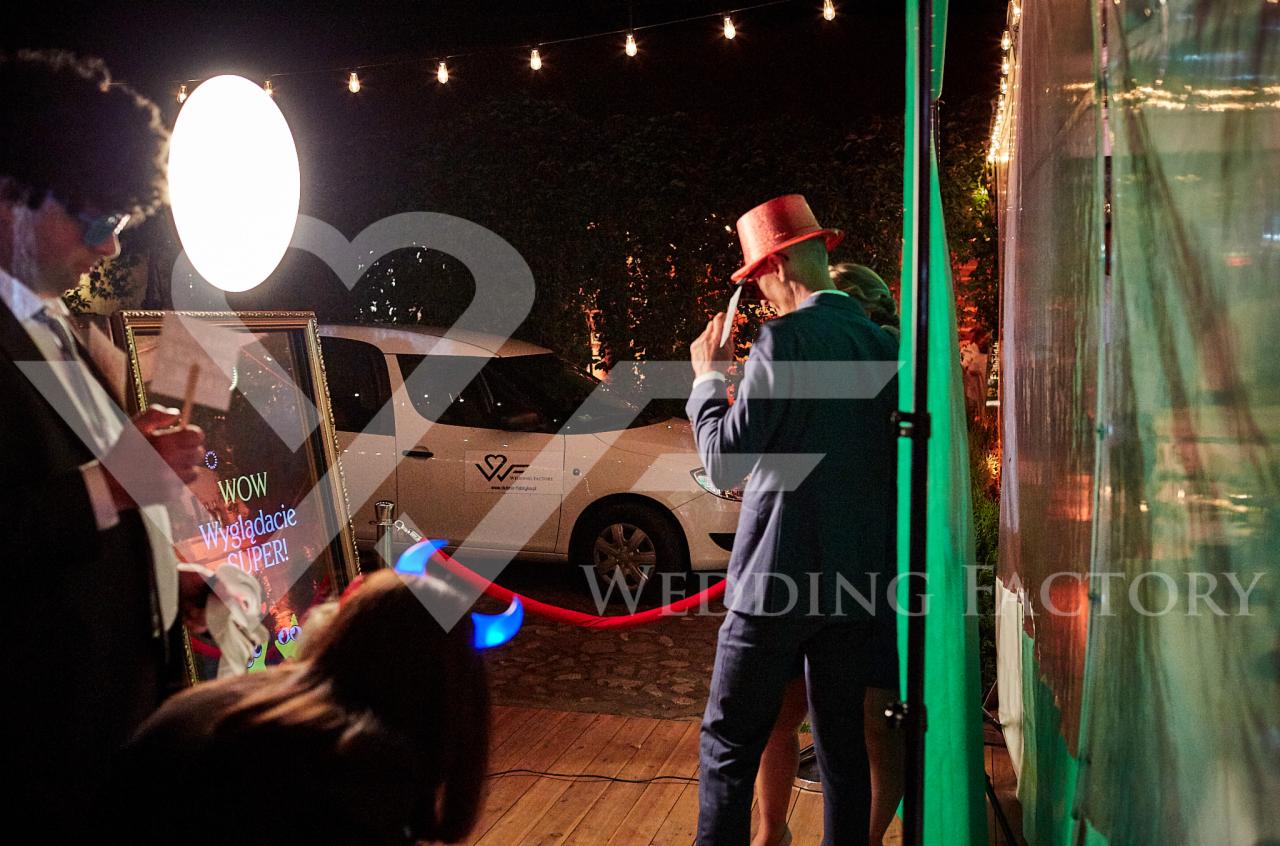 Goście w przebraniach pozują przed lustrem fotobudki z napisem 'WOW Wyglądacie SUPER!' na tle białego samochodu z logo Wedding Factory, oświetlonego girlandą żarówek.