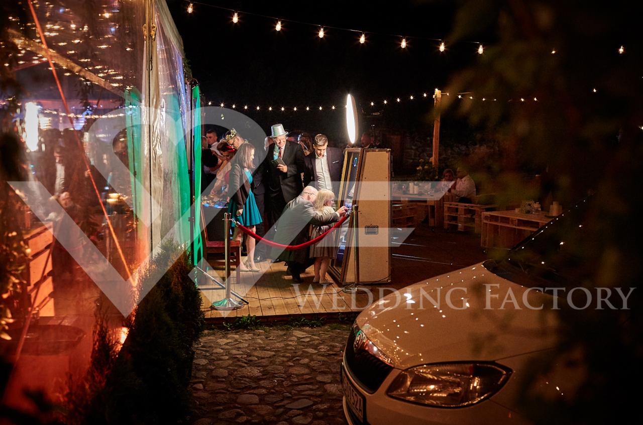 Goście weselni korzystają z fotobudki na zewnątrz w nocy, dekoracja ze światełek, w tle widoczny biały samochód.