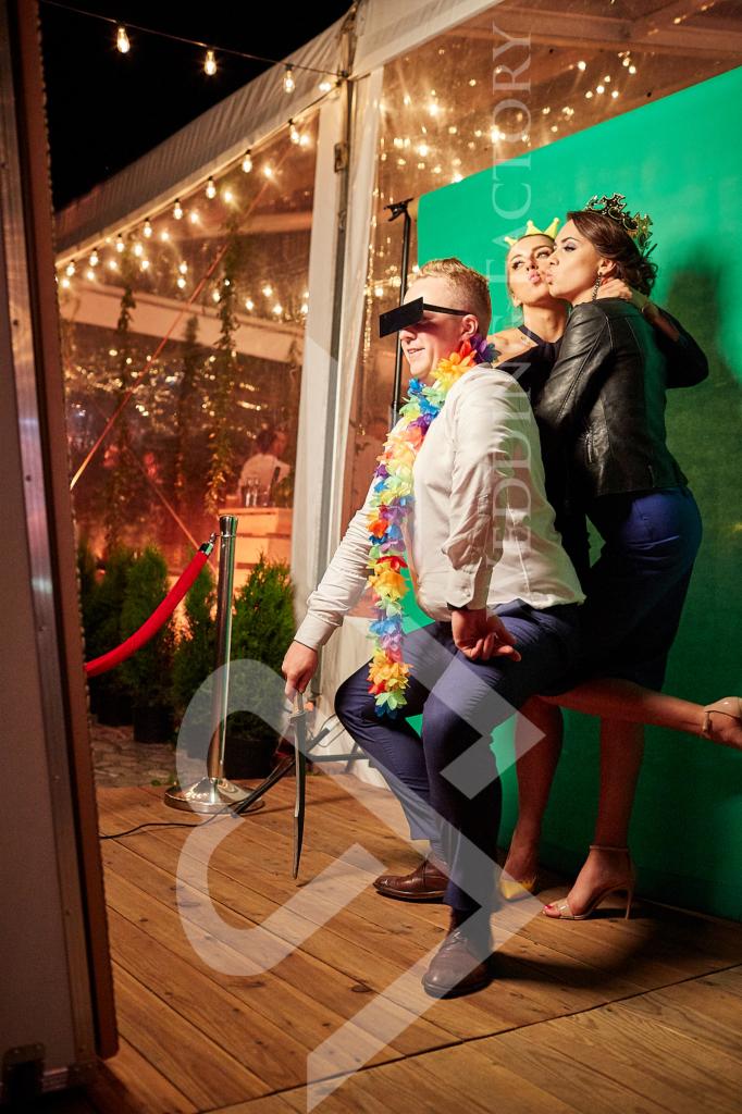 Mężczyzna z czarnymi prostokątnymi okularami i kolorowym naszyjnikiem z kwiatów pozuje z dwiema kobietami w koronie przed zielonym tłem w fotobudce.