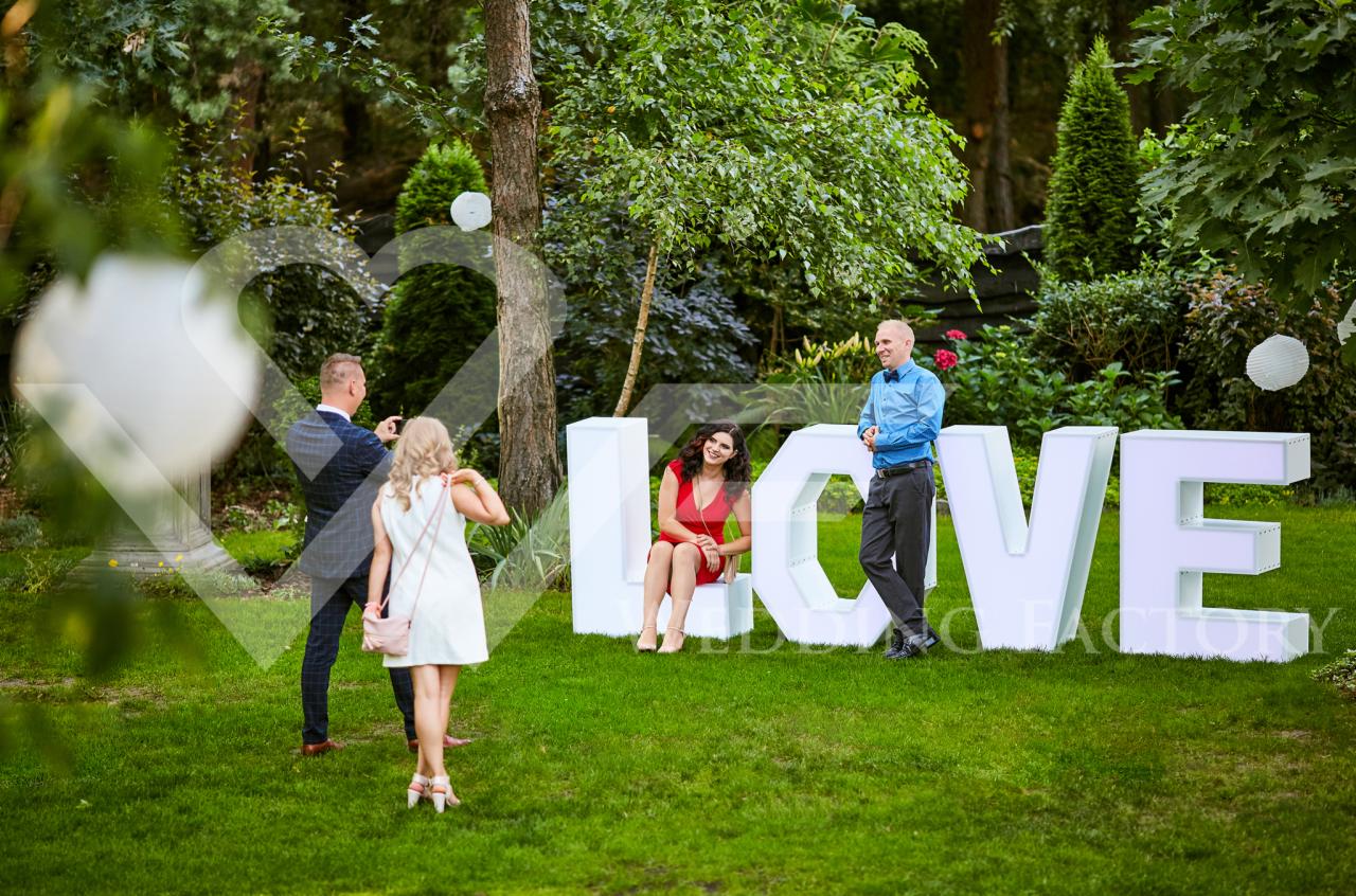 Uśmiechnięta kobieta w czerwonej sukience siedzi na podświetlanym napisie LOVE podczas sesji zdjęciowej w plenerze, fotograf uwiecznia parę w tle.