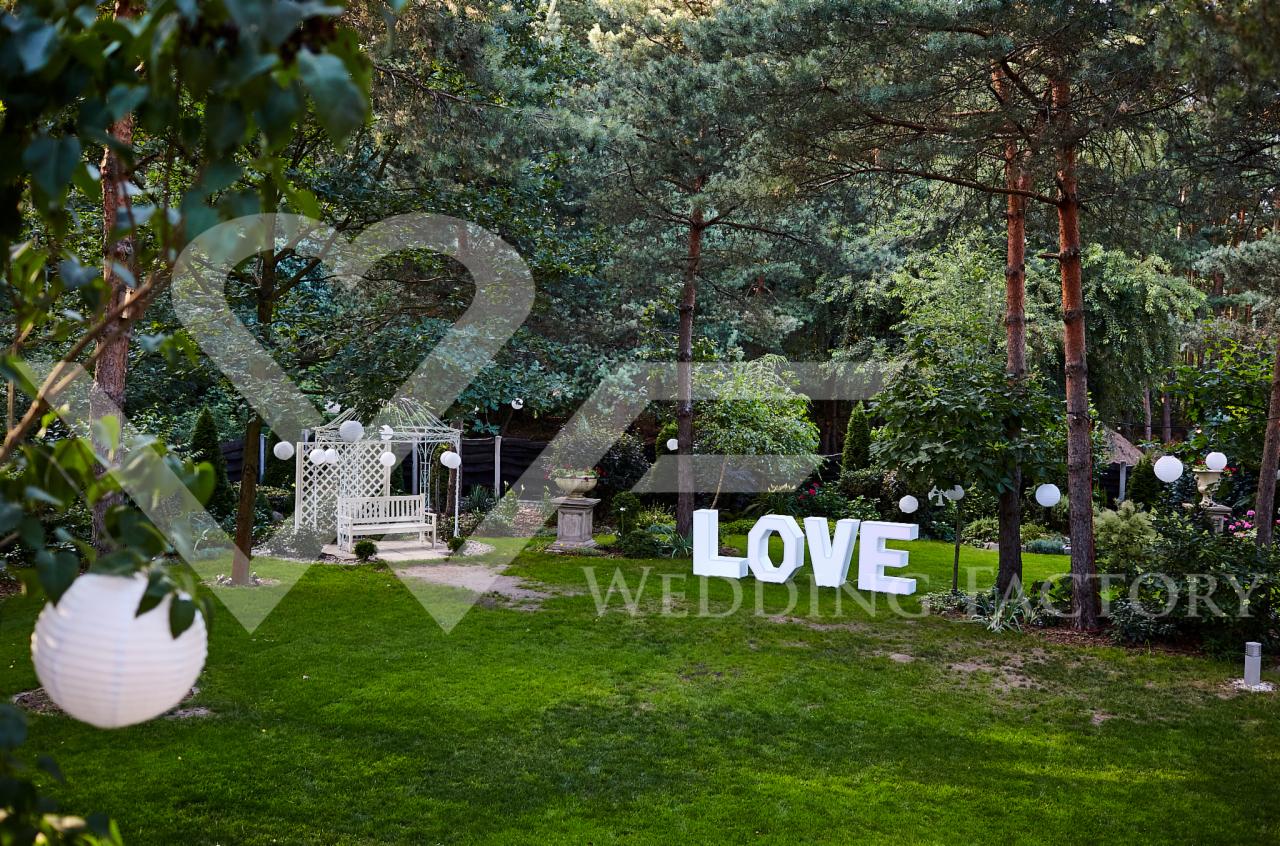 Dekoracja ślubna: biała altana, lampiony i podświetlany napis LOVE WEDDING FACTORY w zielonym ogrodzie.