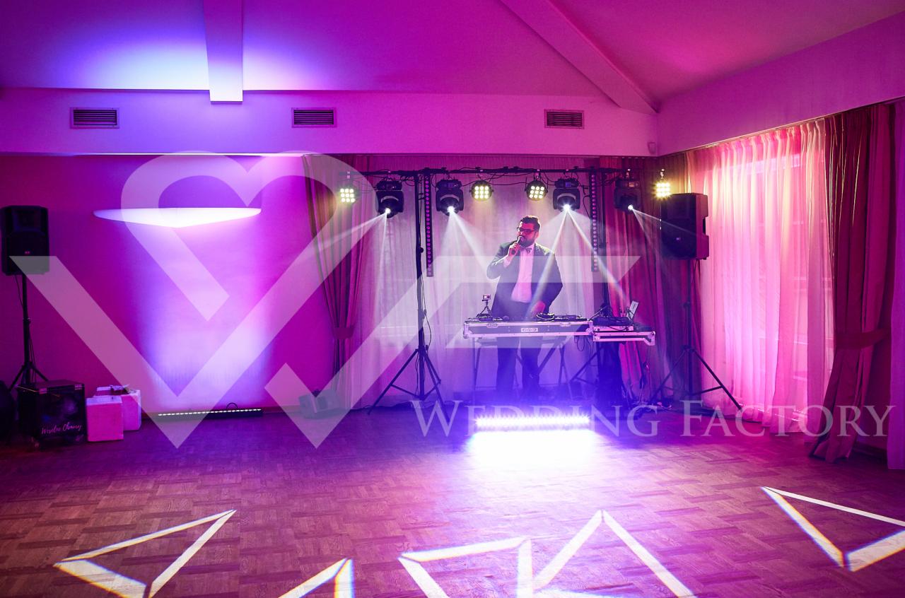 DJ w czarnym garniturze i muszce, z mikrofonem, gra na konsoli DJ-ską na parkiecie z geometrycznymi wzorami świetlnymi, oświetlony reflektorami i fioletowym światłem.