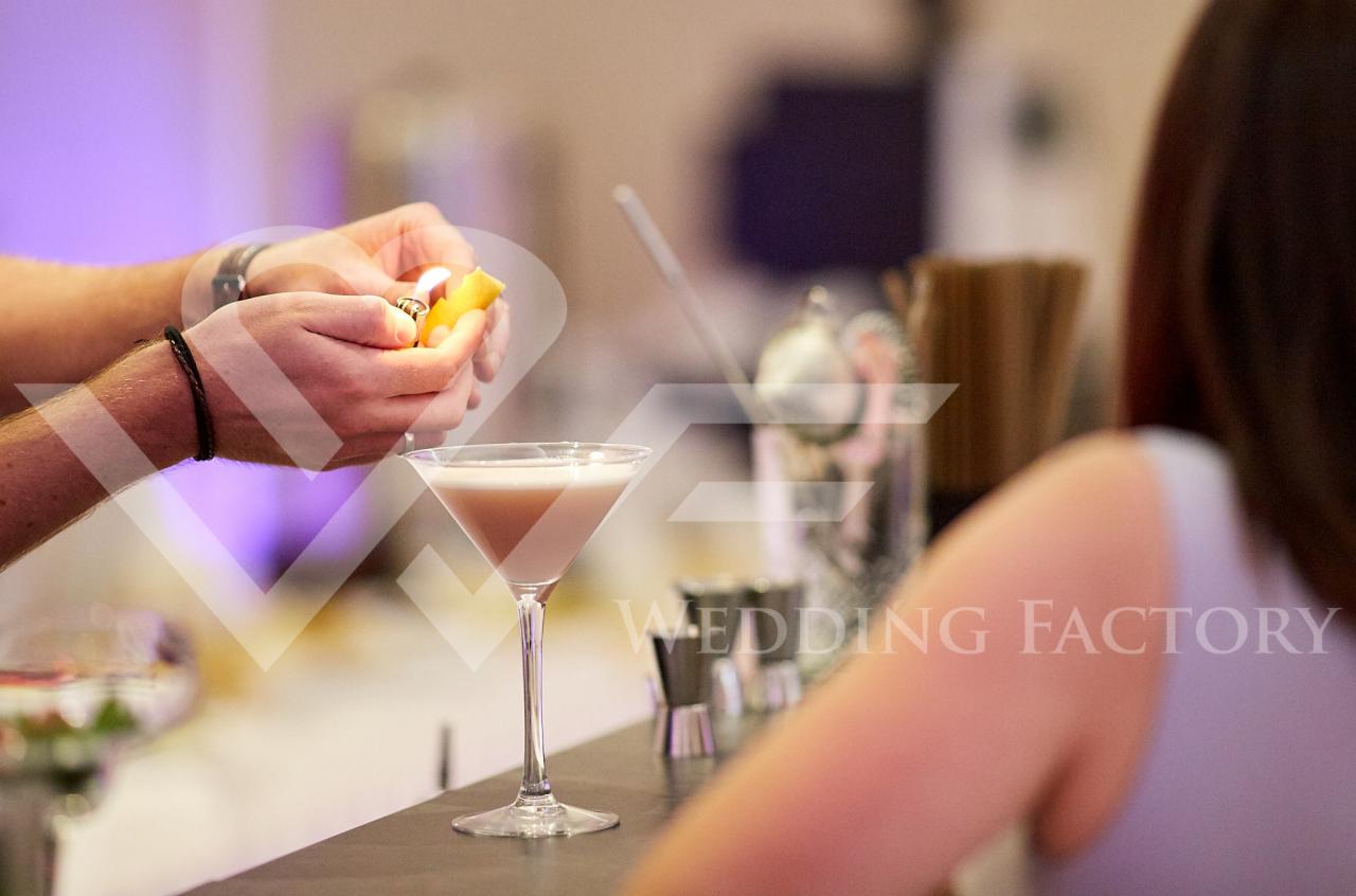 Barman zapala skórkę pomarańczy nad kieliszkiem koktajlu w kształcie litery V, w tle akcesoria barmańskie i fragment sylwetki klienta.