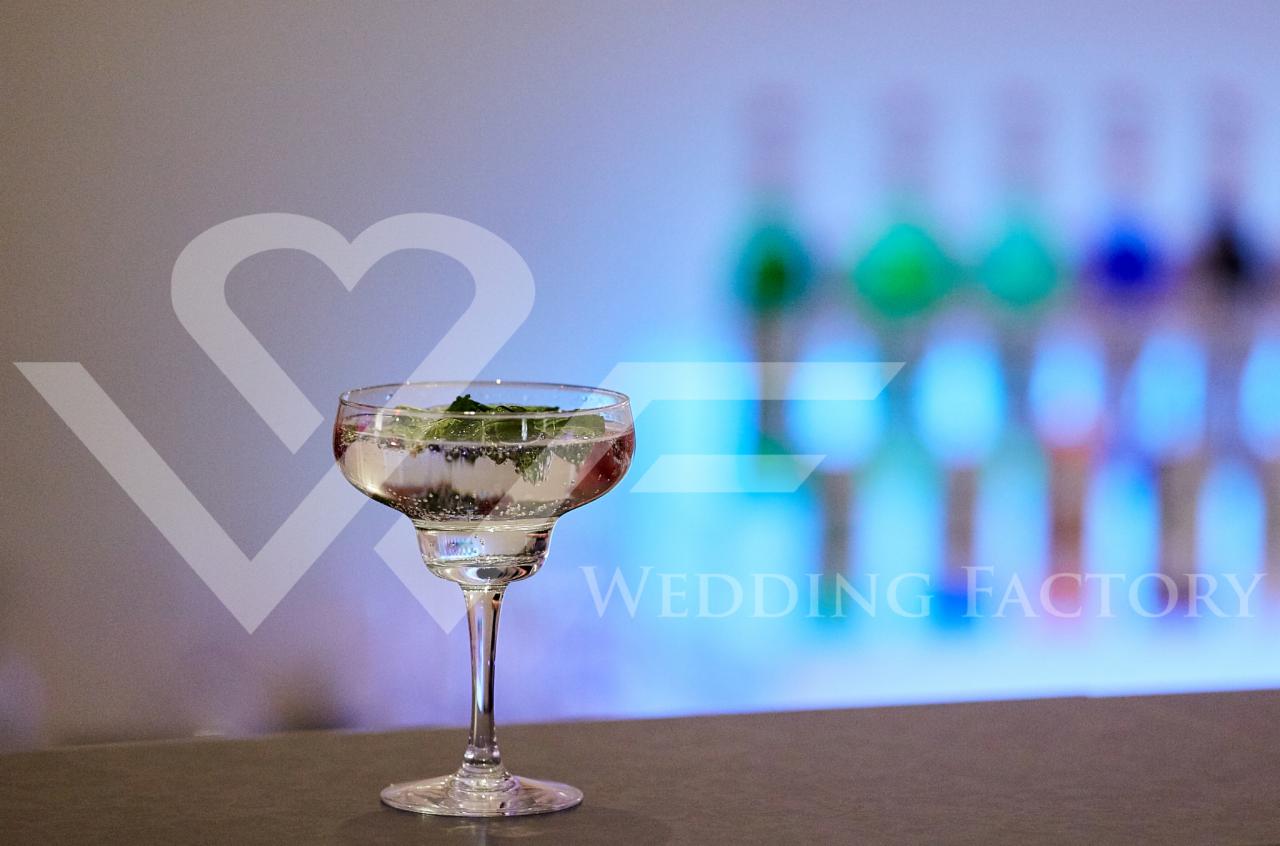 Elegancki kieliszek z gazowanym napojem i listkami mięty na barze, w tle rozmyte butelki i logo Wedding Factory.