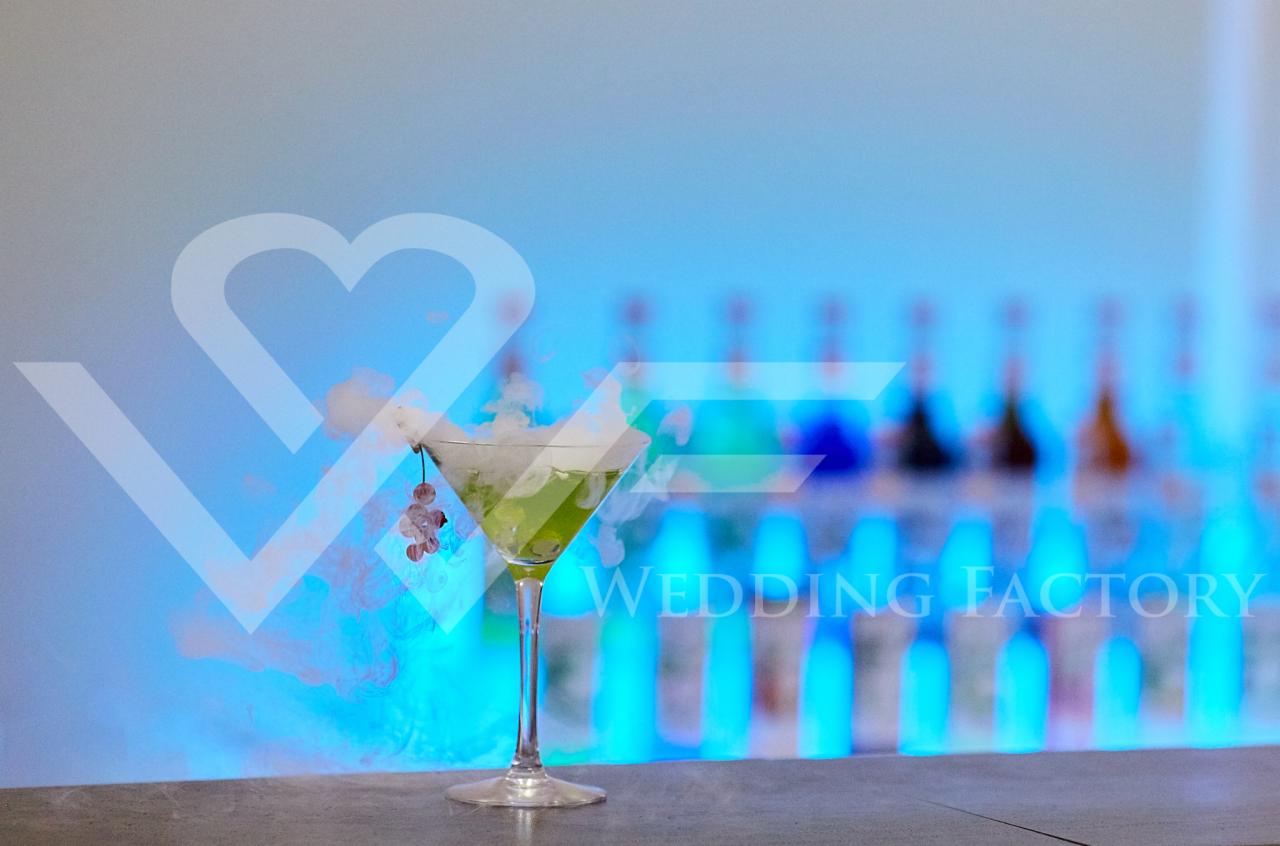 Kieliszek koktajlowy z suchym lodem i dekoracją kwiatową na barze oświetlonym niebieskim światłem, w tle rząd butelek i logo Wedding Factory.