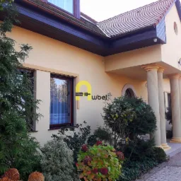 Elewacja domu jednorodzinnego w kolorze kremowym z ciemnobrązową stolarką okienną i kolumnami przy wejściu, widoczna z poziomu ogrodu.