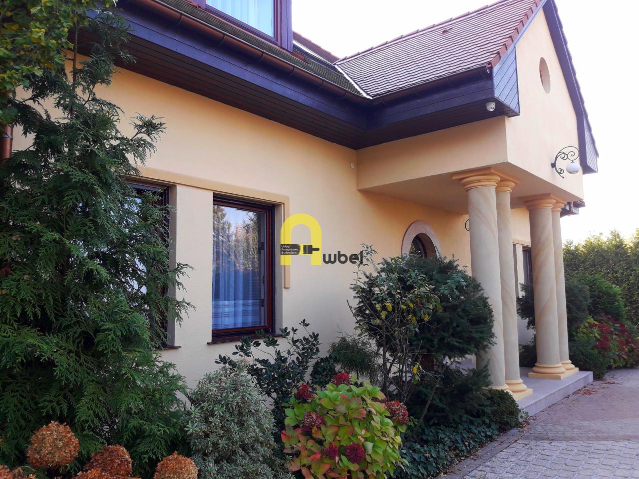 Elewacja domu jednorodzinnego w kolorze kremowym z ciemnobrązową stolarką okienną i kolumnami przy wejściu, widoczna z poziomu ogrodu.