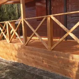 Zadaszony taras drewniany z balustradą w kształcie krzyży, przylegający do brązowego, drewnianego domu z widocznymi drzwiami, z rynną odprowadzającą wodę.