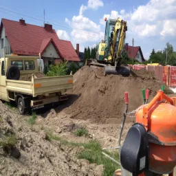 Żółta koparka na usypanej górze ziemi, obok wywrotka z zielonymi krzewami w skrzyni ładunkowej i pomarańczowa betoniarka; w tle domy z czerwonymi dachami i palety cegieł.
