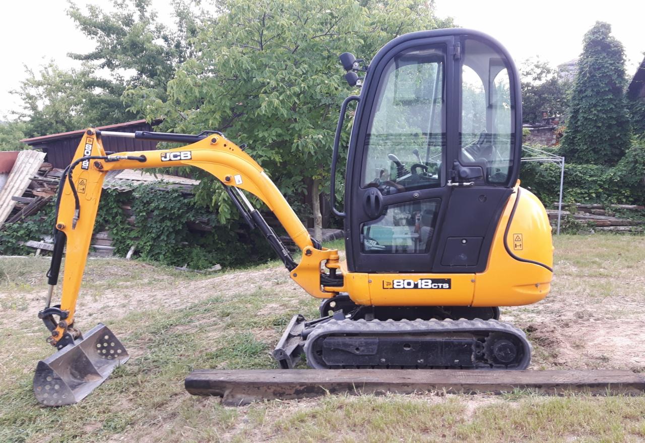 Żółto-czarna mini koparka JCB 8018 CTS zaparkowana na drewnianej belce, widoczna łyżka, gąsienice, kabina operatora, w tle zieleń i zabudowania.