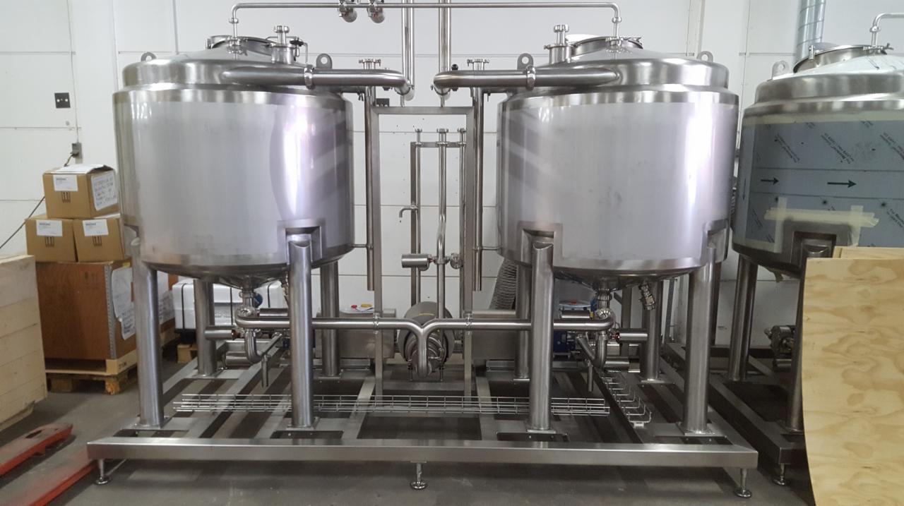 Dwa duże, stalowe zbiorniki na metalowej konstrukcji z rurami, prawdopodobnie w browarze lub zakładzie przetwórstwa spożywczego, widoczne skrzynki i płyty drewniane w tle.