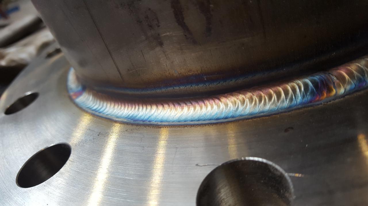 Szczegółowe zbliżenie na spoinę spawalniczą na metalowej flanszy z widocznymi otworami na śruby, ukazujące charakterystyczne przebarwienia termiczne metalu po spawaniu.