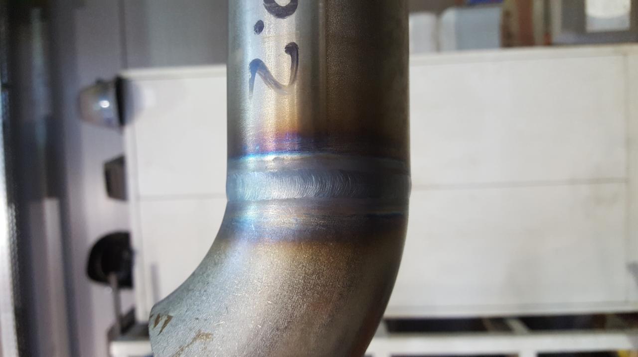 Połączenie spawane rury ze stali nierdzewnej, widoczne ślady po spawaniu i przebarwienia termiczne. Na rurze widoczny napis '2.0'.