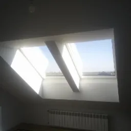 Poddasze z dwoma oknami dachowymi, widok na błękitne niebo i odległy krajobraz, białe ściany i grzejnik pod oknem, skąpane w jasnym, naturalnym świetle.