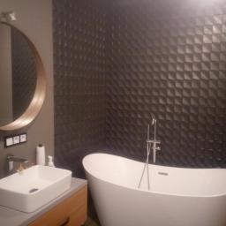 DAR-BUD Dariusz Kucharski - Nowoczesna łazienka z wolnostojącą białą wanną, umywalką nablatową i ścianą wyłożoną ciemnymi, trójwymiarowymi płytkami. Okrągłe lustro z drewnianą ramą zawieszone nad umywalką.