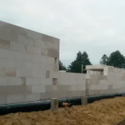 Wznoszenie ścian domu z białych bloczków, widoczne czarne zabezpieczenie fundamentów oraz rury w wykopie, w tle zieleń drzew.