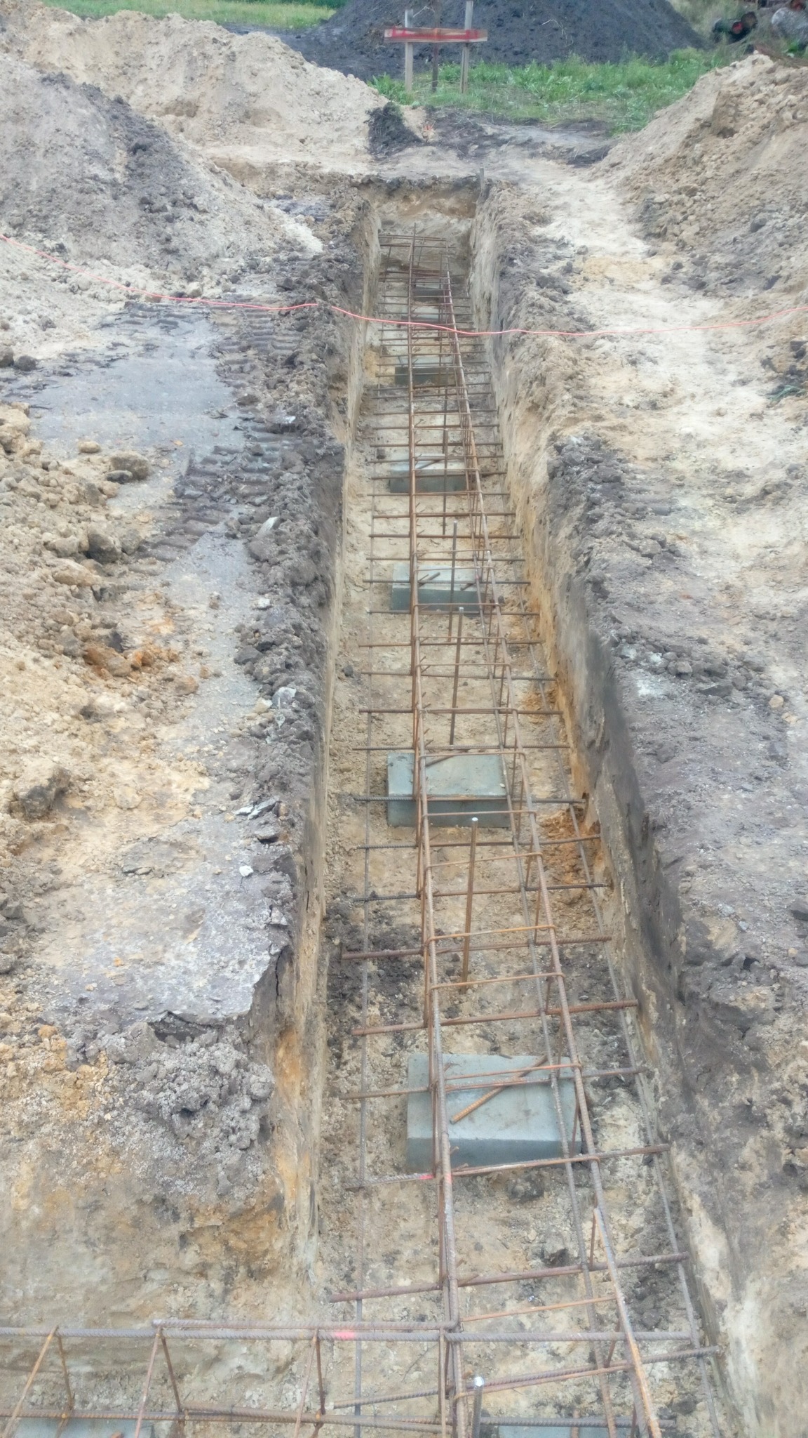 Wykop pod fundament z ułożonym zbrojeniem i betonowymi podkładkami dystansowymi, widoczne ślady po oponach i pryzma ziemi w tle.