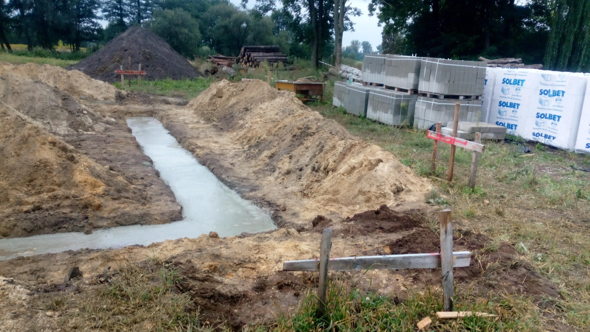 Wykopany fundament zalany świeżym betonem, widoczne sterty ziemi, palety z bloczkami Solbet i stos drewna w tle, teren budowy.