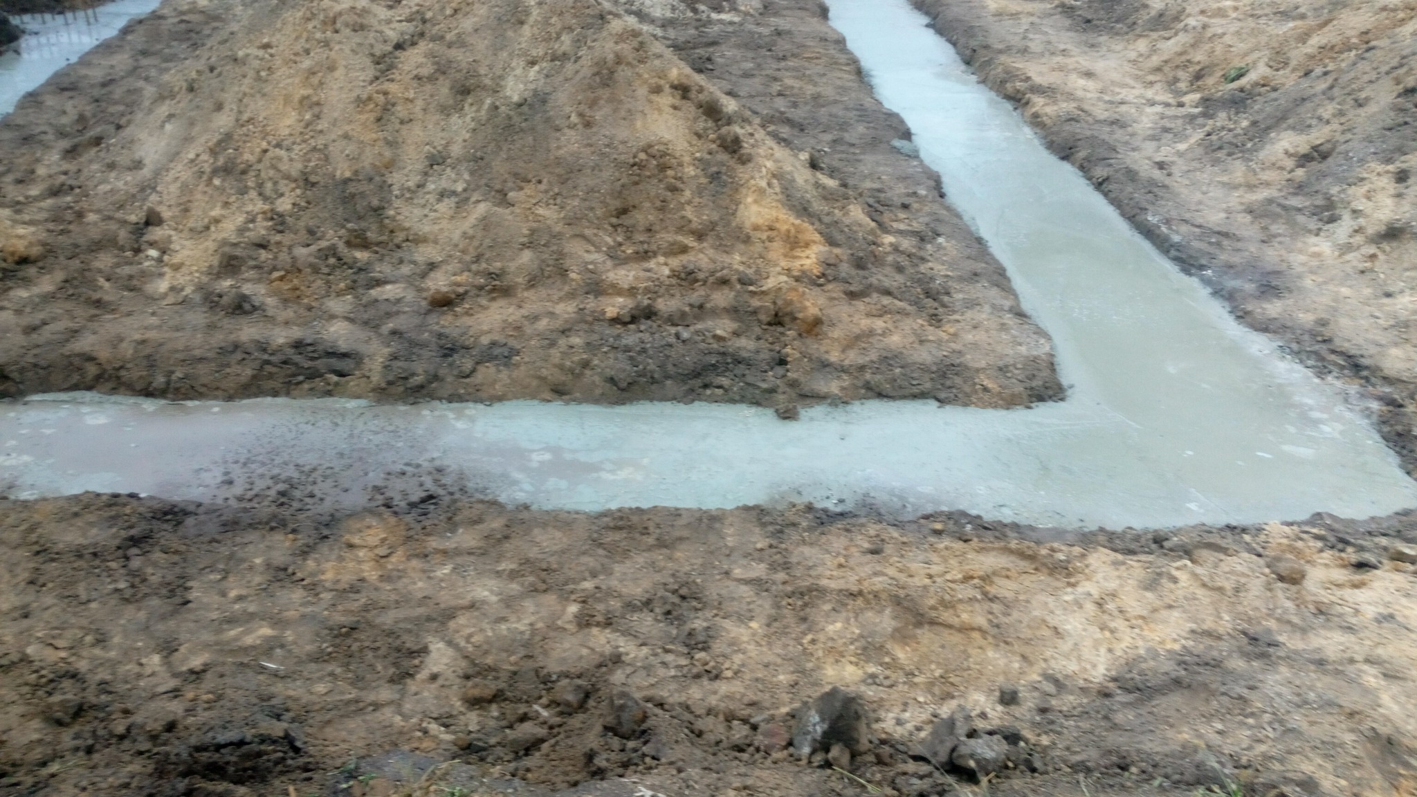 Wykop pod fundament zalany świeżym, szarym betonem, otoczony pryzmami ziemi.