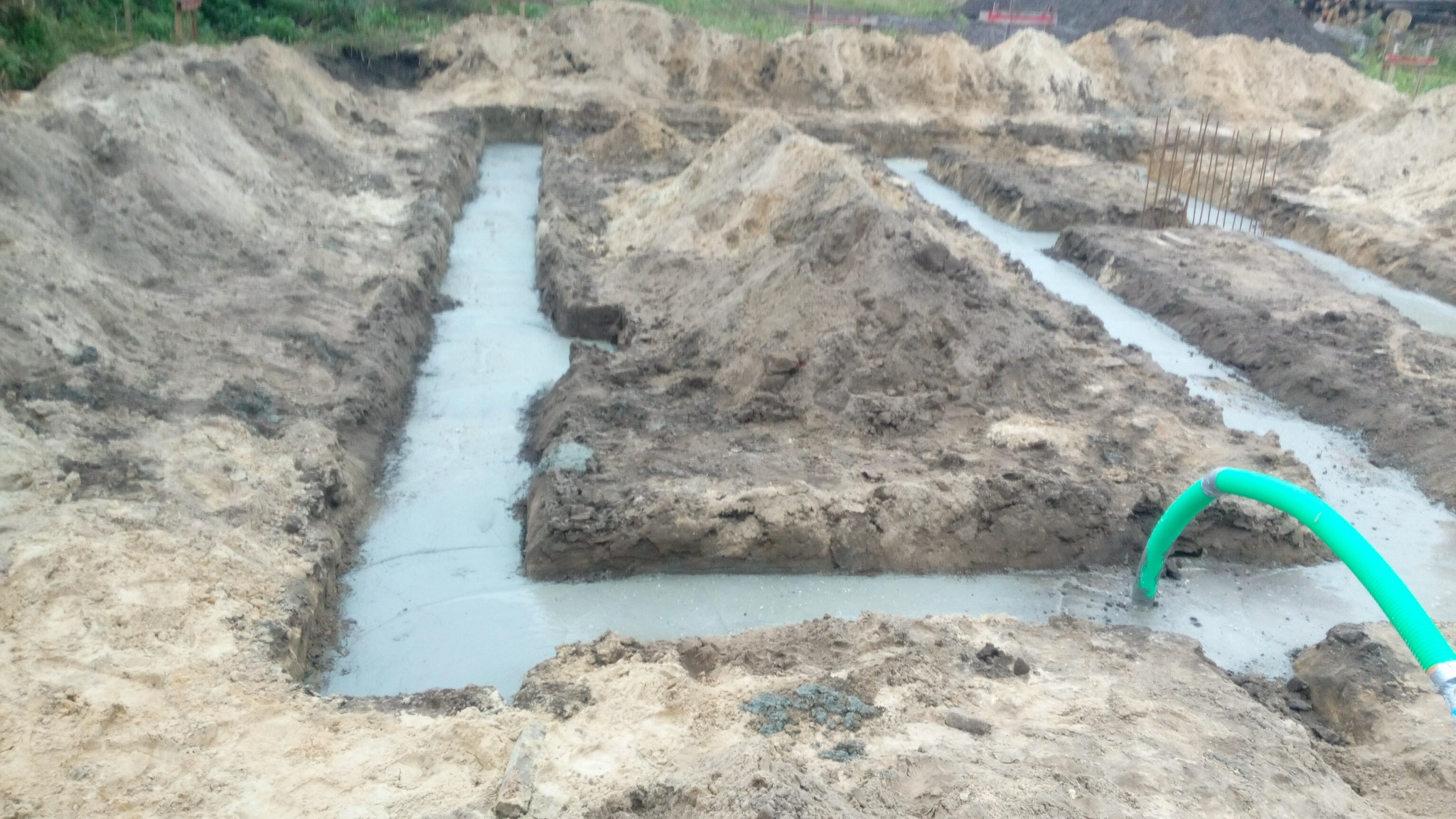 Wylane fundamenty pod budowę domu, widoczne szalunki i świeży beton, zielony wąż doprowadzający wodę do nawilżania betonu.