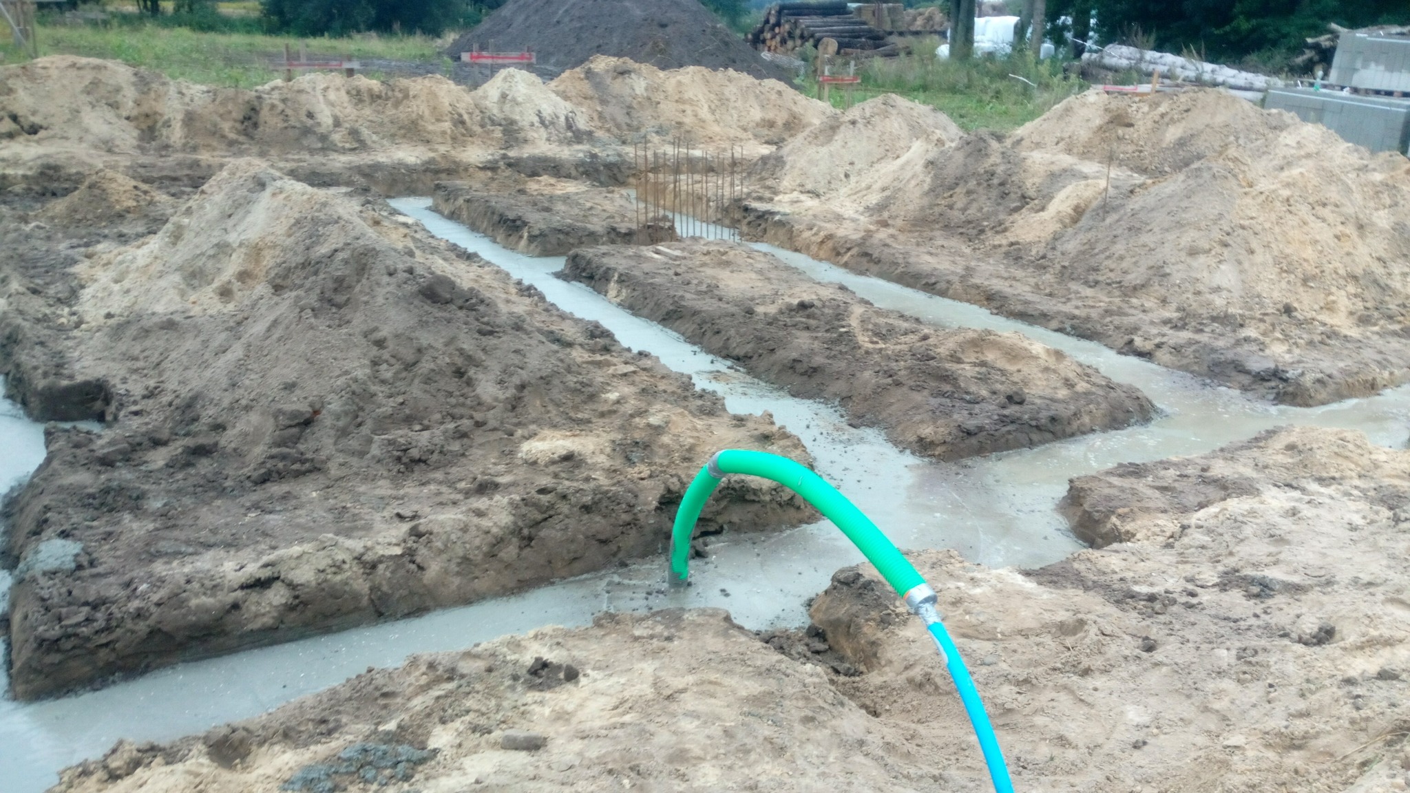 Wykopane rowy fundamentowe wypełnione betonem, z widoczną zbrojeniem i zielonym wężem doprowadzającym wodę, otoczone hałdami ziemi na placu budowy.