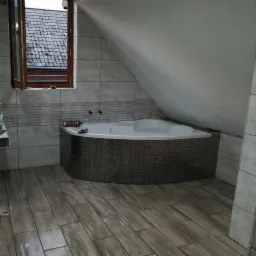 Narożna wanna obudowana mozaiką w łazience na poddaszu, widoczne narzędzia i klej do płytek, drewnopodobna podłoga z płytek.