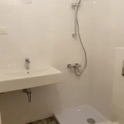 Biała łazienka z umywalką, kabiną prysznicową i toaletą. Na podłodze ciemna mozaika. Ściany wyłożone białymi, błyszczącymi płytkami.