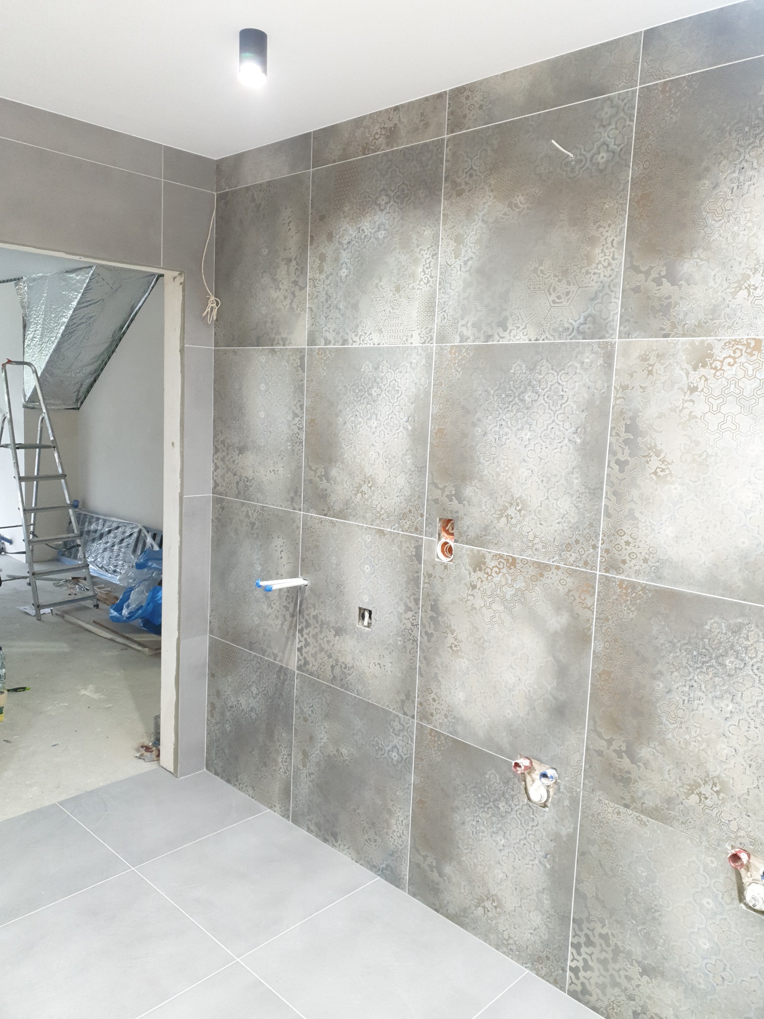 Ściana łazienki wyłożona szarymi płytkami z delikatnym wzorem, widoczne wystające rury instalacji wodnej i elektrycznej, drabina aluminiowa w tle, stan surowy.