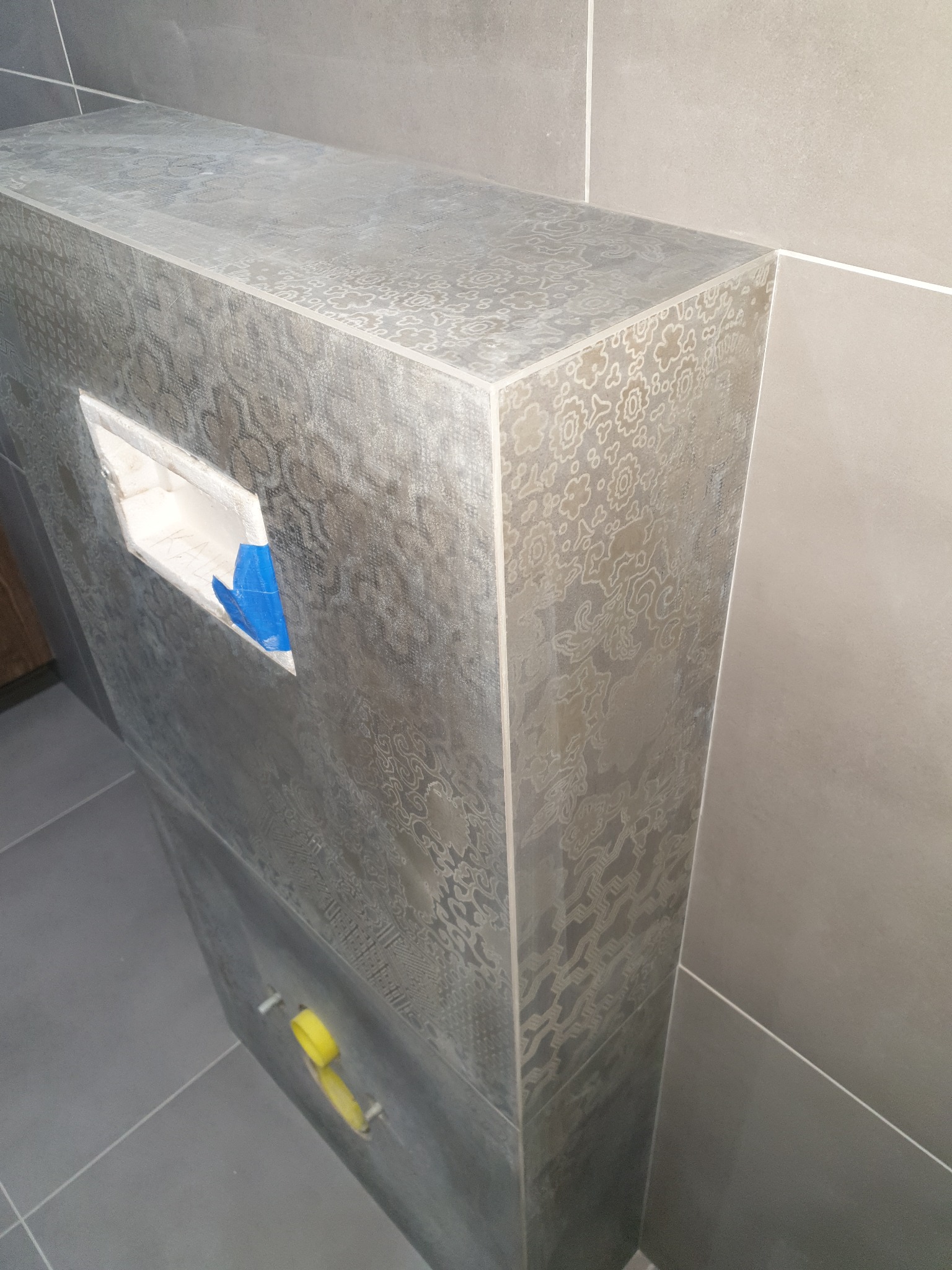 Obudowa WC w łazience wykończona szarą płytką z dekoracyjnym wzorem, widoczna wnęka zabezpieczona niebieską folią i żółta poziomica.