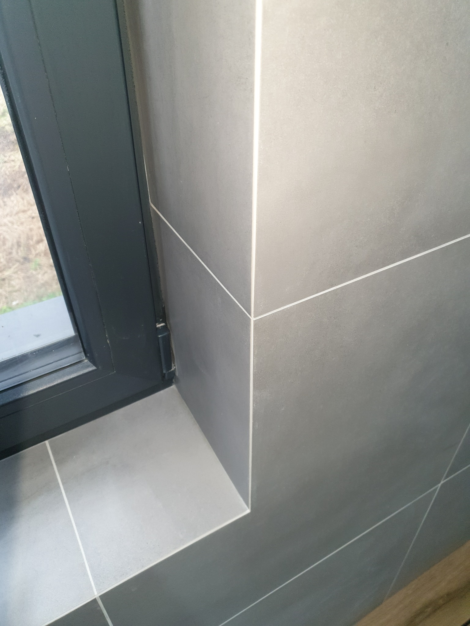 Szare płytki ceramiczne ułożone przy oknie z ciemną ramą, widoczne fugi i krawędź parapetu.