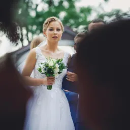 Panna młoda w białej sukni z koronką, trzyma bukiet białych i zielonych kwiatów, otoczona osobami w garniturach, ujęcie z lekkiej perspektywy z rozmazanym pierwszym planem.