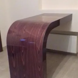 Zaokrąglony blat z ciemnego drewna z widocznym usłojeniem, zamontowany na ścianie w minimalistycznym wnętrzu, z grzejnikiem w tle.