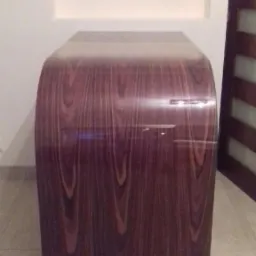 Frontowy widok na nowoczesną szafkę nocną z forniru o ciemnym, bogatym usłojeniu, z zaokrąglonymi krawędziami, stojącą na jasnej podłodze w minimalistycznym wnętrzu.