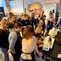 Grupa osób uczestniczy w warsztatach tapicerskich, pracując przy maszynach do szycia. W tle widoczny baner z logo 'AK Design Akademia Rzemiosła' oraz elementy dekoracyjne przypominające skrzydła.