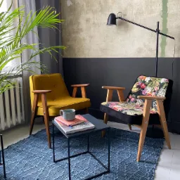 Dwa odnowione fotele w stylu retro: jeden żółty, drugi z tapicerką w kwiaty, stoją na niebieskim dywanie obok stolika kawowego z książkami i kubkiem, na tle ściany w dwóch odcieniach.