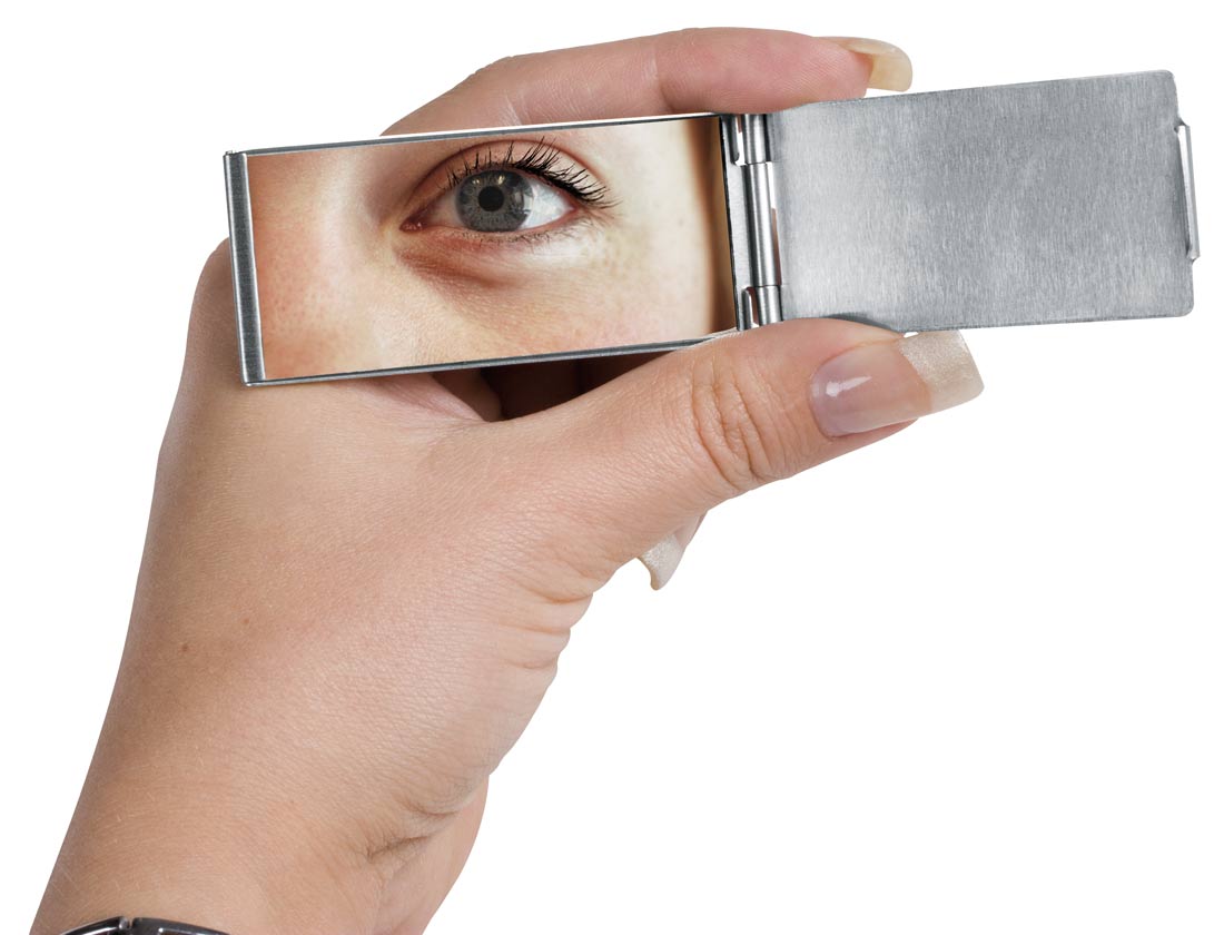 Otwarty, prostokątny, metalowy lusterko kieszonkowe, trzymane w dłoni, odzwierciedla zbliżenie na szaro-zielone oko kobiety z widocznymi piegami i drobnymi zmarszczkami.