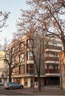 Nowoczesny, trzypiętrowy budynek mieszkalny z balkonami, widoczny z poziomu ulicy, otoczony drzewami z nagimi gałęziami, zaparkowany szary samochód osobowy przed budynkiem.