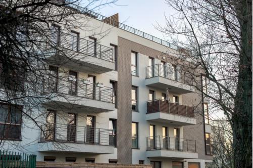 Nowoczesny budynek mieszkalny z balkonami, częściowo zasłonięty przez gałęzie drzew bez liści, w jesiennej scenerii.