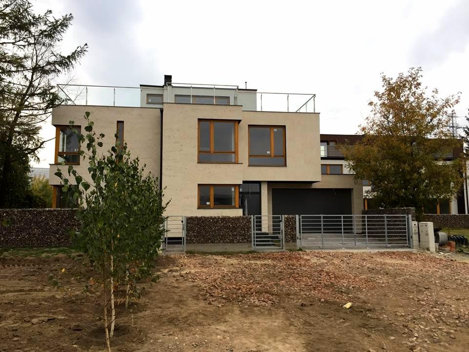 Nowoczesny, dwukondygnacyjny dom jednorodzinny z płaskim dachem i szklaną balustradą na dachu, ogrodzony metalową bramą i gabionami, z młodymi drzewkami na pierwszym planie i ziemią po robotach...