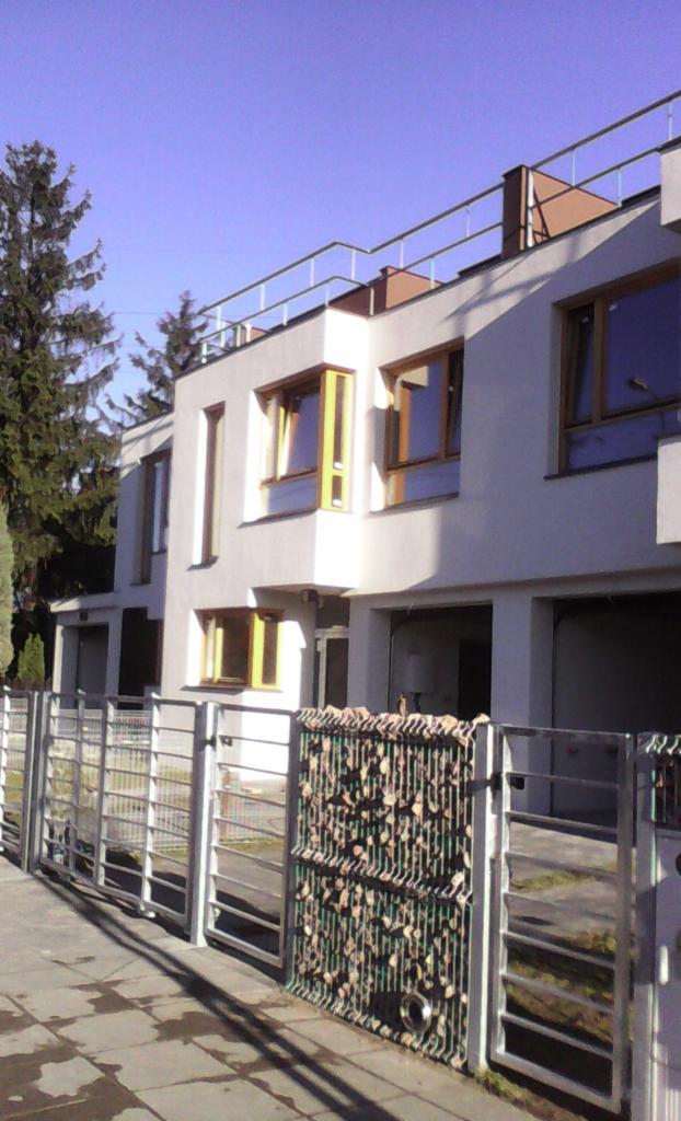 Nowoczesny, dwupiętrowy budynek z płaskim dachem i metalową balustradą, widziany zza metalowego ogrodzenia z elementami dekoracyjnymi z kamieni i siatki.