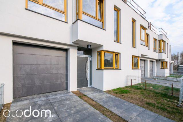 Nowoczesny szeregowiec z białymi elewacjami, szarą bramą garażową i żółtymi ramami okiennymi, widok z zewnątrz.