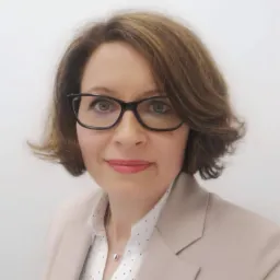 Portret kobiety w okularach, w beżowej marynarce i bluzce w kropki. Subtelny makijaż i krótka, brązowa fryzura. Jasne tło.