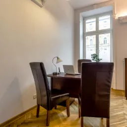 Biuro z drewnianym parkietem w jodełkę, biurkiem, laptopem, lampką i trzema krzesłami z brązowej skóry, widok na okno z dekoracyjnymi sztukateriami.