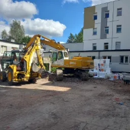 Dwa żółte, ciężkie maszyny budowlane na placu budowy, z gruzem po rozbiórce w tle i stosem materiałów izolacyjnych oraz fragmentem budynku wielorodzinnego w tle.