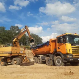 Żółta koparka CASE CONTEC ładuje piasek do pomarańczowej ciężarówki Scania 114C 380 na piaszczystym terenie pod błękitnym niebem z chmurami.