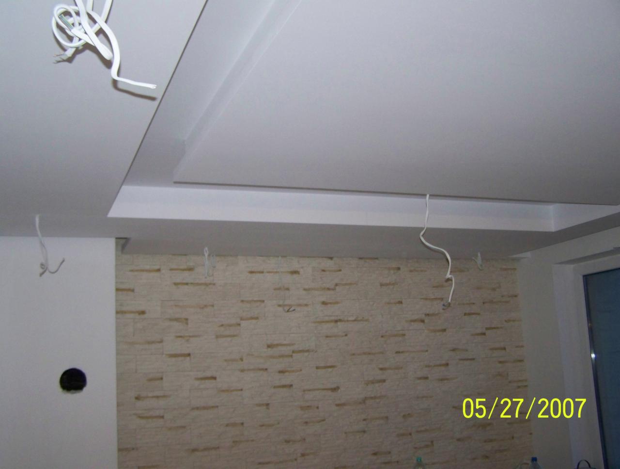 Sufit podwieszany z oświetleniem w trakcie montażu, widoczne wiszące kable, ściana wyłożona dekoracyjną cegłą w odcieniach beżu.