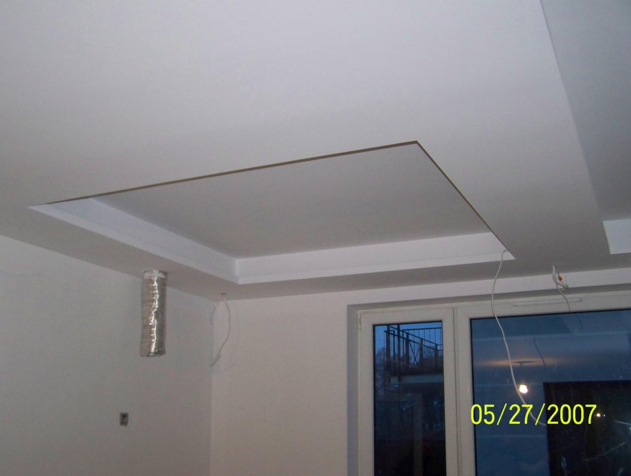 Biały sufit podwieszany z kwadratową wnęką i listwami dekoracyjnymi, widoczny przewód wentylacyjny i okno z odbiciem balkonu.