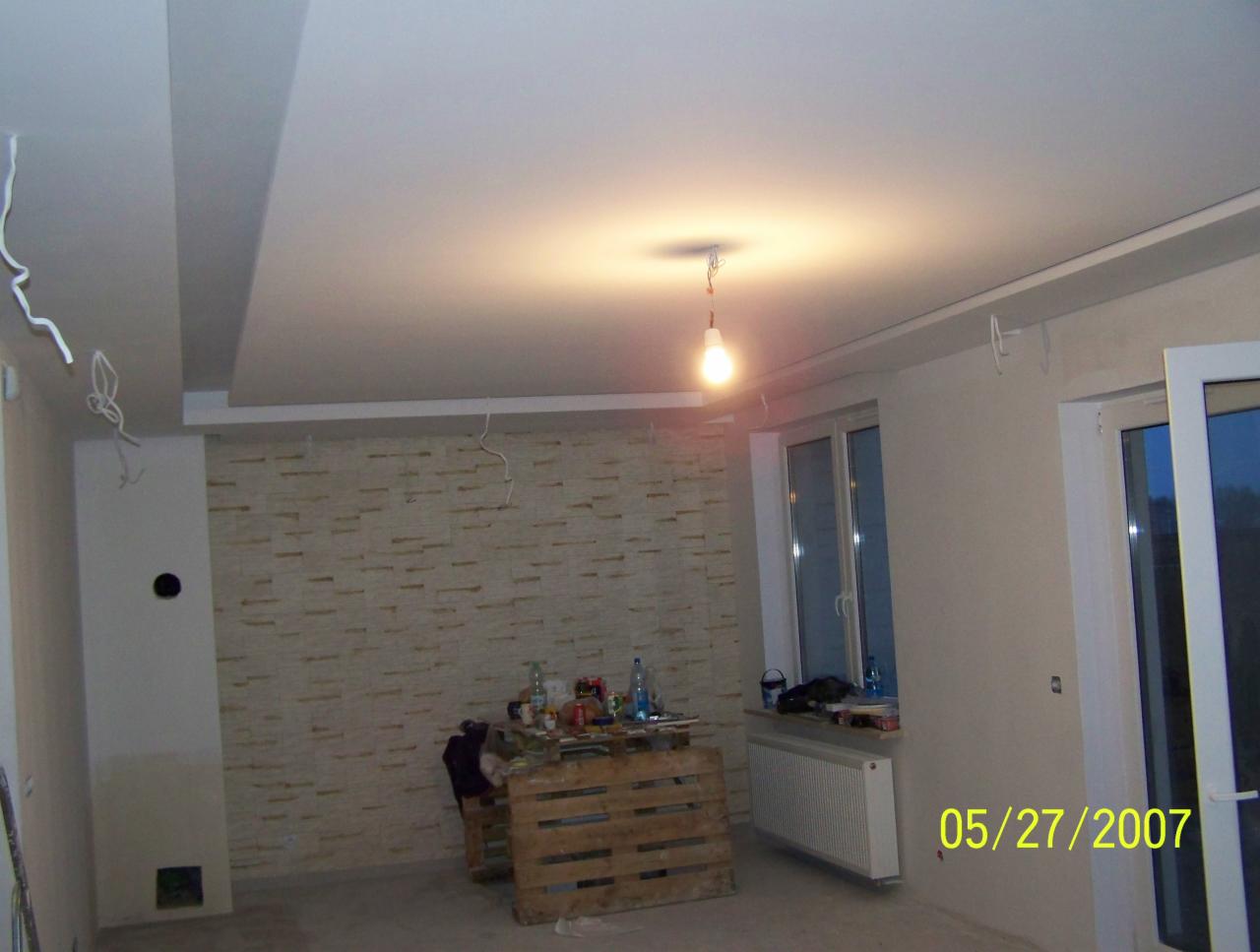 Pokój w trakcie prac wykończeniowych: położona częściowo dekoracyjna okładzina ścienna z imitacji kamienia, podwieszany sufit z wystającymi kablami, prowizoryczne oświetlenie żarówką i okno...