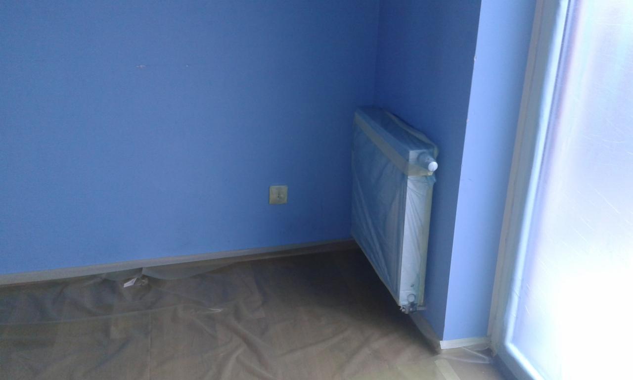 Świeżo pomalowane na niebiesko pomieszczenie z grzejnikiem zabezpieczonym folią malarską i podłogą przykrytą folią ochronną, widoczny gniazdko elektryczne na ścianie.