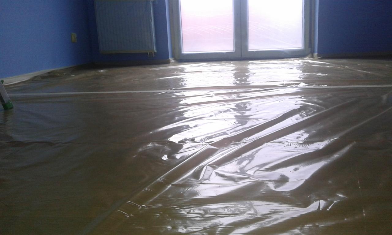 Podłoga w pomieszczeniu zabezpieczona folią malarską przed remontem, widoczne ściany w kolorze niebieskim, grzejnik i okno zabezpieczone folią.